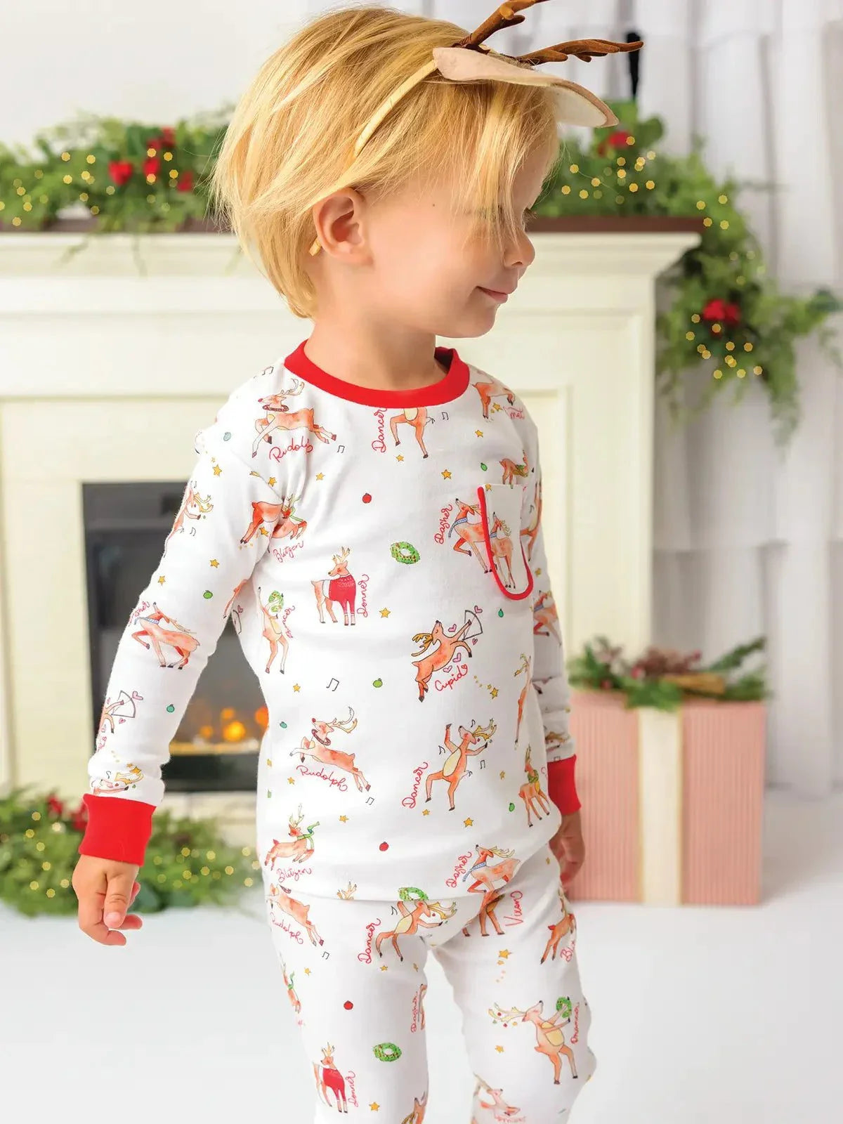 Child wearing organic cotton Christmas pajama set with reindeer print and red cuffs in festive holiday room