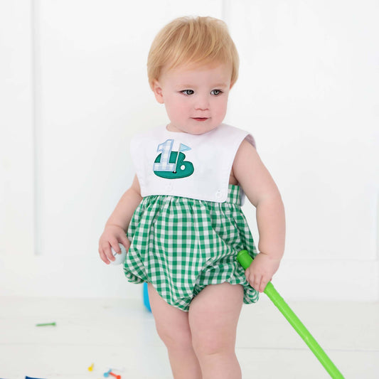 Toddler boy wearing Hole-In-One First Birthday reversible golf bubble outfit with green checkered shorts and white bib.